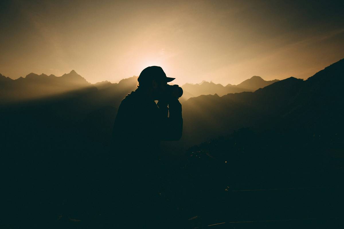 A hiker holding a DSLR camera while standing on a scenic mountaintop overlooking valleys below.