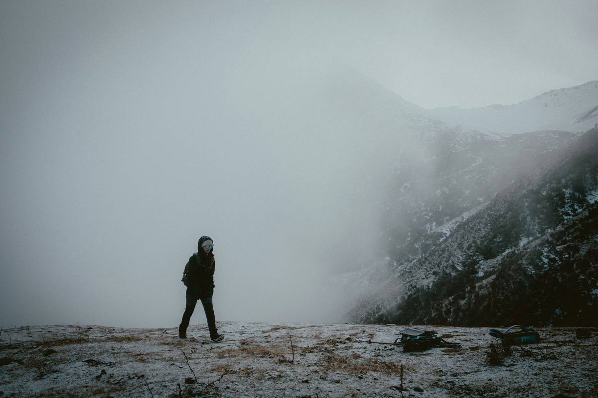 A hiker adjusting a small portable camera attached to their backpack, surrounded by lush greenery and sunlight filtering through trees.
