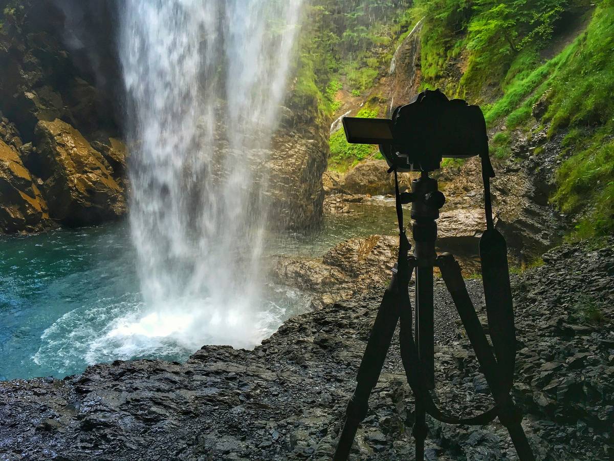 A lightweight mirrorless camera attached to a backpack with a tripod standing nearby on a rocky trail.